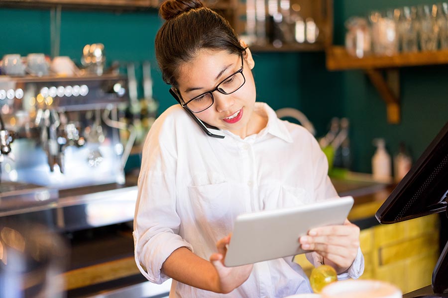 Contact Us - Restaurant Owner Uses a Tablet and Cell Phone Behind the Counter, Wearing Glasses and a White Dress Shirt