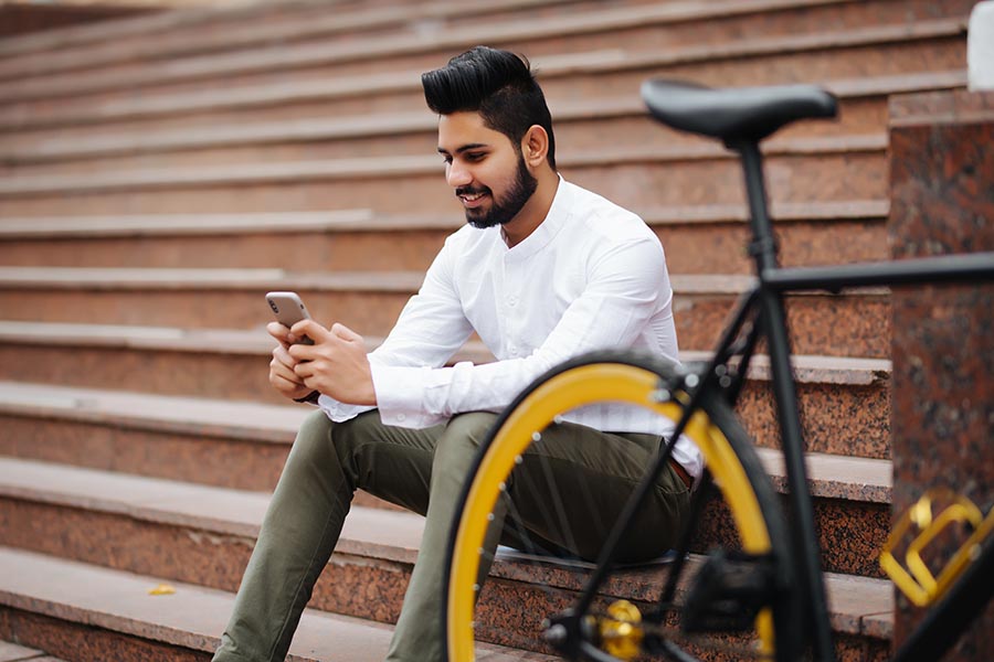 Mobile App - Young, Well-Dressed Man Uses Smartphone Sitting on Public Stairs With His Bike Next to Him